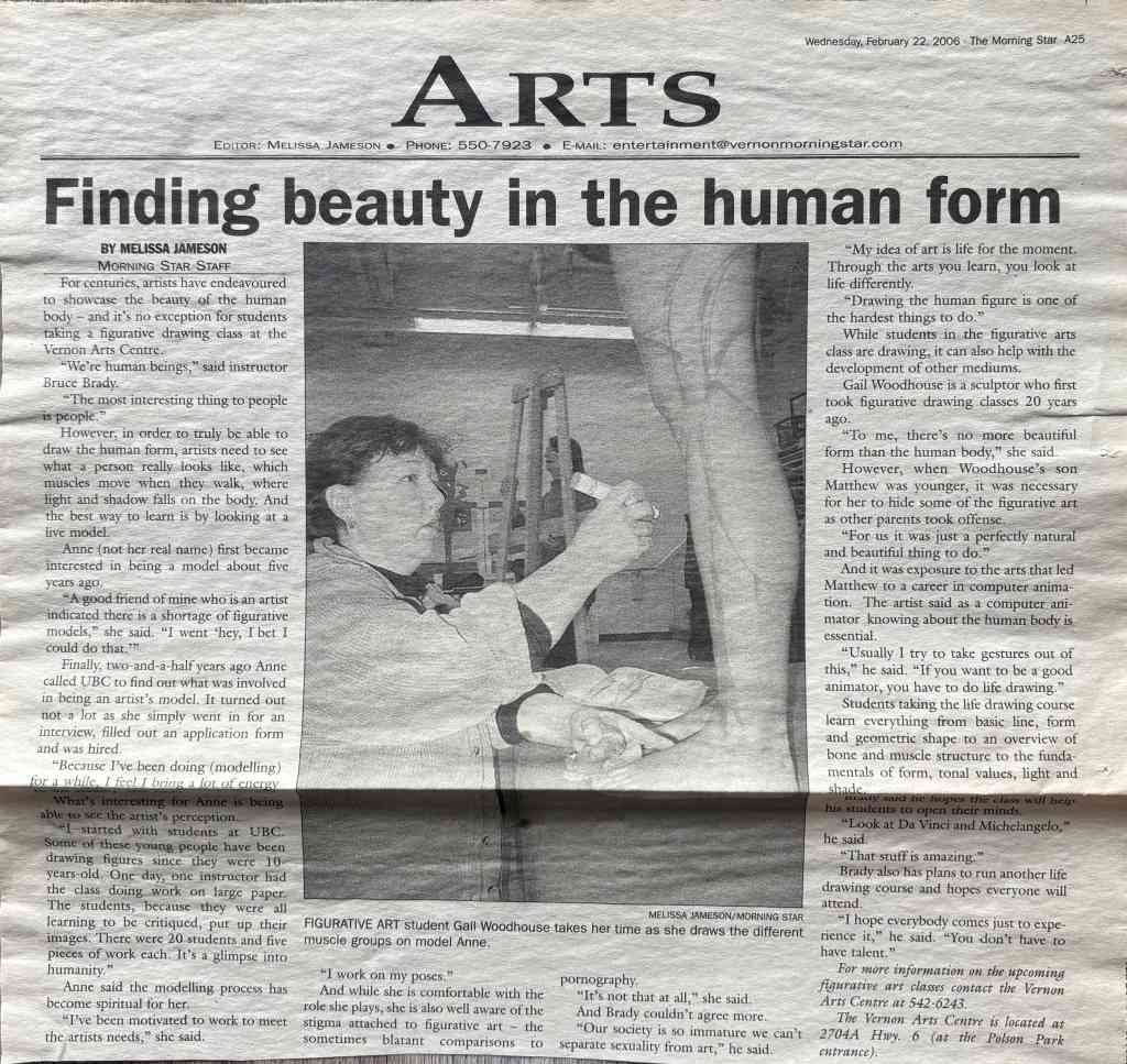 Black-and-white photo of a person seated at a table, holding up a sculpted head or mask toward a camera; studio tools and artworks are visible in the background.